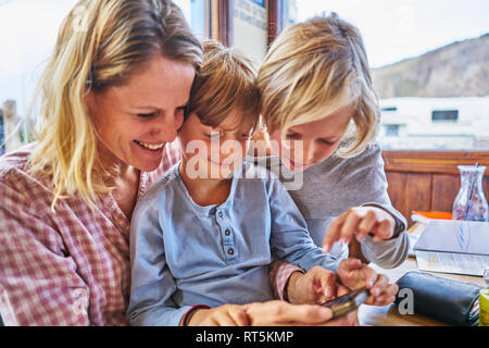 Heureuse fête avec deux fils assis à table dans un café à jouer avec cell phone Banque D'Images