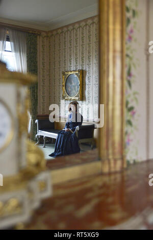 Une vieille femme se distingue par un style Victorien très cheminée décorée et regarde dans un miroir- il y a une horloge bronze doré d'or disant que c'est 4h00 à côté d'elle. Banque D'Images