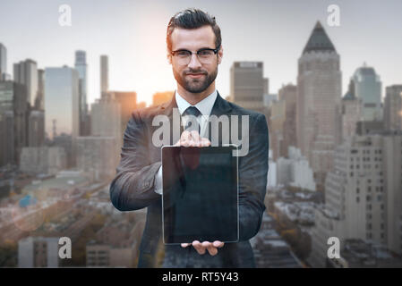 Jetez un coup d'oeil ! Portrait of young man in suit showing digital tablet at camera while standing against de matin cityscape background. Concept numérique. Smart Business. Technologies Banque D'Images