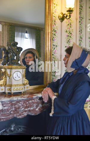 Une vieille femme se distingue par un style Victorien très cheminée décorée et regarde dans un miroir- il y a une horloge bronze doré d'or disant que c'est 4h00 à côté d'elle. Banque D'Images