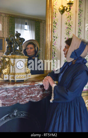 Une vieille femme se distingue par un style Victorien très cheminée décorée et regarde dans un miroir- il y a une horloge bronze doré d'or disant que c'est 4h00 à côté d'elle. Banque D'Images