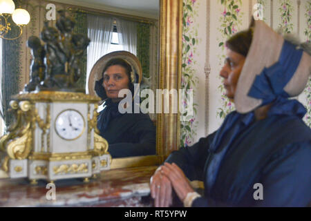 Une vieille femme se distingue par un style Victorien très cheminée décorée et regarde dans un miroir- il y a une horloge bronze doré d'or disant que c'est 4h00 à côté d'elle. Banque D'Images