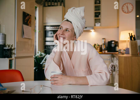 Portrait of smiling woman wearing towel turban assis avec une tasse de café à la table dans la cuisine Banque D'Images