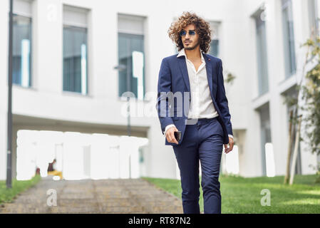 Portrait de jeune homme à la mode avec des cheveux bouclés portant des lunettes de soleil et costume bleu Banque D'Images