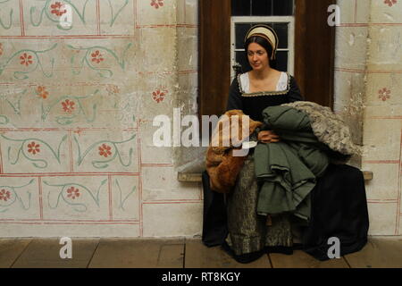 Une belle jeune femme en robe est Tudor authentique en appareil photo et sourit- elle est assise dans une vieille fenêtre et tient les vêtements.
