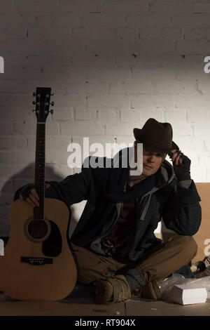 Sans-abri dans la région de hat assis sur carton par white brick wall et holding guitar Banque D'Images
