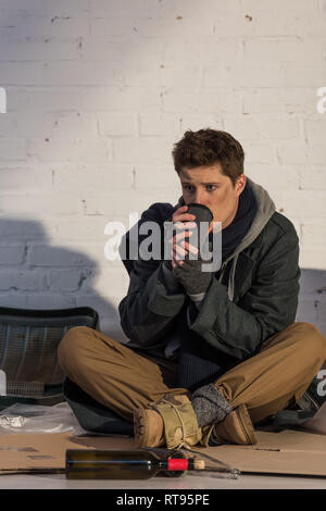 Triste homme sans foyer d'alcool et de tasse de papier alors qu'il était assis sur le mur de brique en carton Banque D'Images