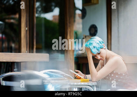 Jeune femme pensive avec les cheveux teints en bleu assis dans un café de la chaussée à l'écoute de la musique avec des écouteurs Banque D'Images
