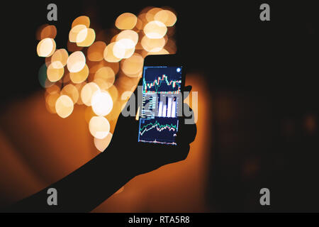 Woman hand holding mobile phone avec stock trading sur l'écran dans l'obscurité avec la lumière d'arrière-plan flou flou, financières, du commerce, des affaires et de techno Banque D'Images