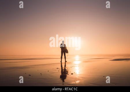 A man holding a surfboard, silhouetté contre le soleil couchant, Saunton, Devon, UK. Banque D'Images