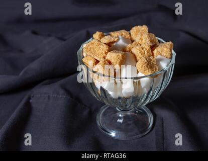 Cubes de sucre brun et blanc dans un vase en verre sur un fond de tissu foncé. Draperie. Focus sélectif. Image rectangulaire. Banque D'Images