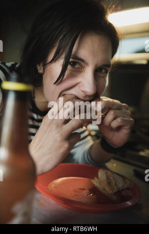 Femme assise à table, mordre près de sandwich plat et bouteille de verre et à la recherche à l'appareil photo en mobile home Banque D'Images