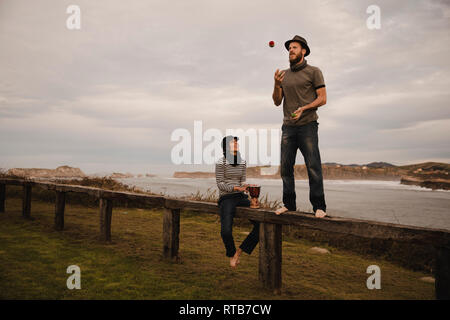 Jeune mec en hat juggling balls près de dame élégante en cap avec tambour d'éthique s'asseoir sur le siège près de la côte de mer et ciel nuageux Banque D'Images