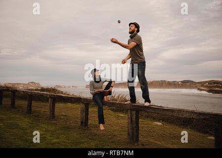 Jeune mec en hat juggling balls près de dame élégante en cap avec tambour d'éthique s'asseoir sur le siège près de la côte de mer et ciel nuageux Banque D'Images