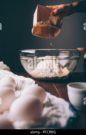 Close-up of bowl pour pétrir la pâte avec des tas de farine et le beurre en dés à côté des oeufs crus. Woman cooking gâteau fait maison sur table en bois brun, vertical Banque D'Images