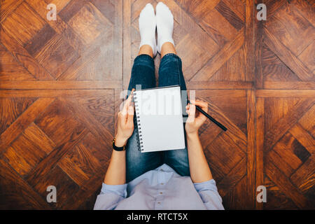 Top View of young woman sitting at home sur plancher en bois holding blank notebook. Banque D'Images