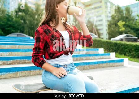 Jeune fille lycéenne adolescent 10-15 ans, summer city après les leçons à l'école. Il est assis sur une planche à roulettes, dans ses mains smartphone, tasse de café ou de boissons Banque D'Images