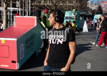 Jeune homme portant des écouteurs dans un T-Shirt noir avec slogan Brixton Hun marche à côté de 'presque' 3D signe à Kings Cross, London Banque D'Images