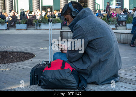 Jeune homme sittting et portant un casque à la mode et à la recherche à son mobile près de la gare de Kings Cross, Londres, Angleterre Banque D'Images