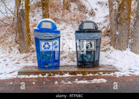 Poubelles avec pente enneigée et arbres arrière-plan. Une piscine bleu et noir poubelle placée sur le côté de la route. Une pente couverte de neige avec des arbres c Banque D'Images