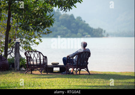 Triste vieillard dans une veste assis sur une chaise sur une plage de lonely et regardant la mer Banque D'Images