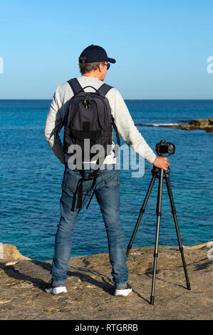 Vue arrière d'un photographe portant un sac à dos et debout par une caméra et trépied face à la mer. Banque D'Images