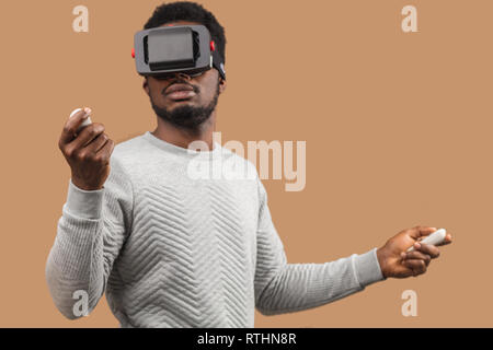 Studio shot of African American man à l'aide de casque de réalité virtuelle, de l'exploitation avec la manette en mains, de se déplacer sur un fond beige Banque D'Images