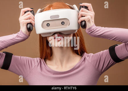 Gingembre occasionnels-haired woman en violet pullower portant casque VR et à impliqués dans le jeu de simulation 3D, désignant les mains comme s'il en interaction avec Banque D'Images