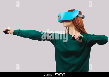 Cheerful young female with ginger hair maintenant si elle handsas visant de la proue, explorer la réalité virtuelle alors qu'il se trouvait isolé sur gray backgroun Banque D'Images