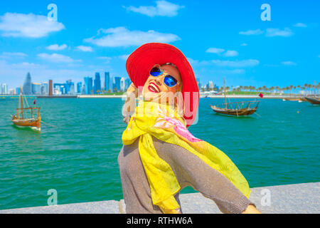 Femme heureuse avec chapeau le long de la baie de Doha avec un boutre traditionnel vieux bateaux en arrière-plan. Portrait style touriste jouit d'gratte-ciel de Doha Banque D'Images