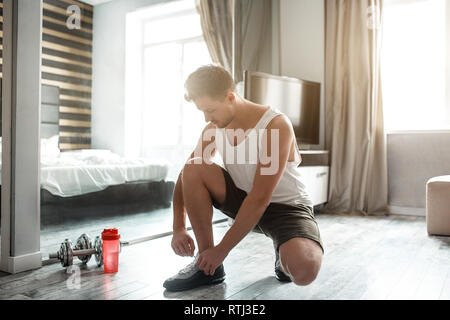 Jeune homme bien bâti du sport dans l'appartement. Guy s'asseoir sur un genou et les lacets sur les chaussures de sport. Bouteille d'eau rouge se placer à côté de l'haltère. Dayligh Banque D'Images