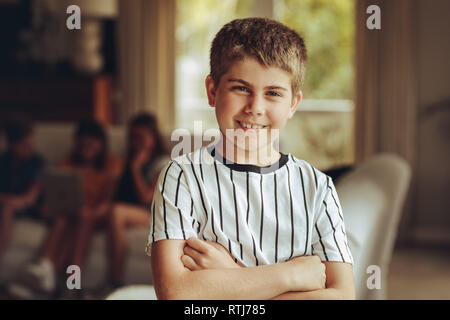 Close up d'un innocent à la garçon debout avec les mains pliées. Portrait d'un garçon debout à la maison avec ses amis assis en arrière-plan. Banque D'Images