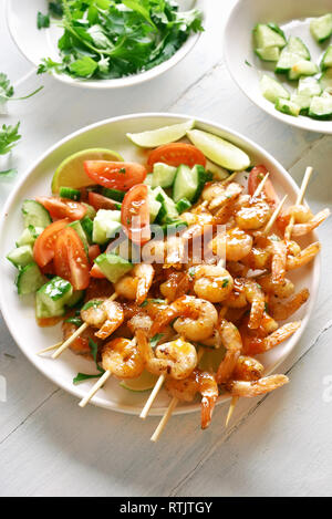 Brochettes de crevettes grillées sur salade de légumes et de tranches de concombre et les tomates sur la plaque blanche sur table en bois. Brochettes de crevettes grillées avec une sauce Banque D'Images