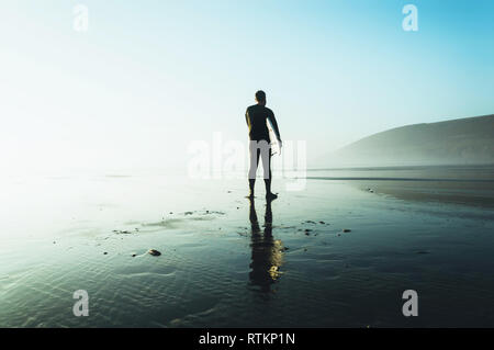 Un internaute tenant un surf silhouetté contre le soleil d'après-midi, sur un hivers brumeux jour. Saunton, Devon, UK. Banque D'Images