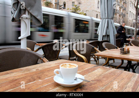 Une tasse de café se trouve sur une table de café en plein air tandis que le tramway de Jérusalem passe par dans un flou en arrière-plan. Banque D'Images