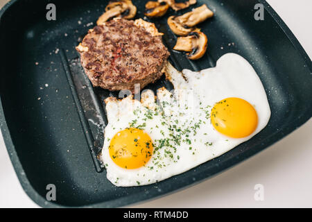 Au-dessus de la viande de boeuf cru délicieux burger cuit dans une poêle à frire sur la surface en teflon grill cuisine avec deux oeufs au plat avec champignons frits organique Banque D'Images