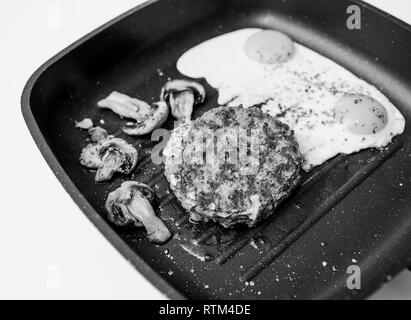 Au-dessus de la viande de boeuf cru délicieux burger cuit dans une poêle à frire sur la surface en teflon grill cuisine avec deux oeufs au plat avec champignons frits organique image en noir et blanc Banque D'Images