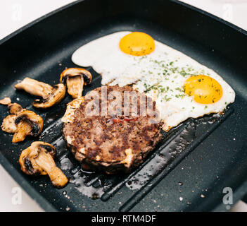 Au-dessus de la viande de boeuf cru délicieux burger cuit dans une poêle à frire sur la surface en teflon grill cuisine avec deux oeufs au plat avec champignons frits organique Banque D'Images