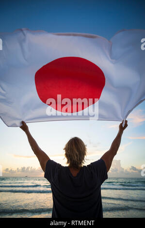 Sunrise pittoresque silhouette of man holding drapeau japonais en face de soft ciel du matin au-dessus d'une plage avec des vagues qui arrive sur l'horizon Banque D'Images