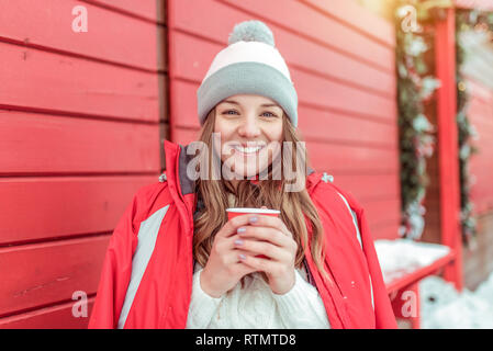 Belle femme en hiver dans une veste rouge sur fond de volets en bois. Tenant une tasse de café de thé. Heureux sourire et rire. Maison de vacances resort Banque D'Images