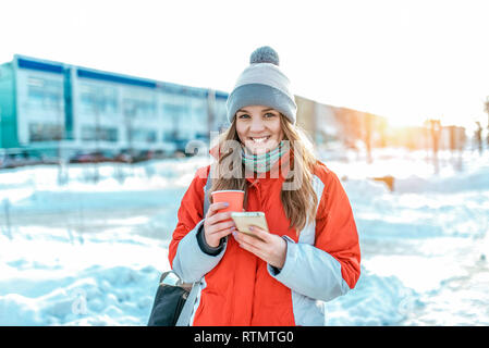 Femme heureuse est debout sur la rue en hiver en ville. Dans sa main une tasse de café ou de thé. Compose un message sur téléphone. Les émotions de bonheur, de sourires et de Banque D'Images