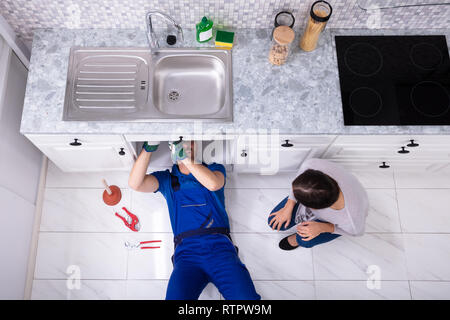 High Angle View of Woman Lying On Floor Handyman Repairing Sink In Kitchen Banque D'Images