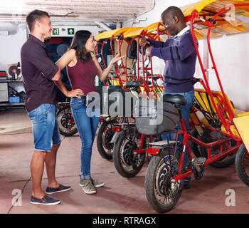 L'homme afro-américain de pilote insatisfait parle de pousse-pousse avec couple de jeunes clients agréable Banque D'Images