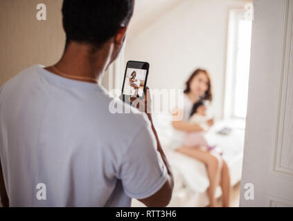 Attractiave jeune mère jouant avec son bébé pendant que papa prend des photos d'eux à la maison.fond flou, se concentrer sur la photo dans le smartphone Banque D'Images