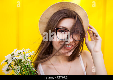 Hipster girl heureux portant des lunettes et un chapeau avec des fleurs à l'arrière-plan jaune. Tenue d'été. CLose up of smiling woman Banque D'Images