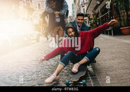 Souriante jeune femme est poussée sur planche par son petit ami à l'extérieur sur rue, avec des amis sur le dos en arrière-plan dans la rue. Groupe de fr Banque D'Images