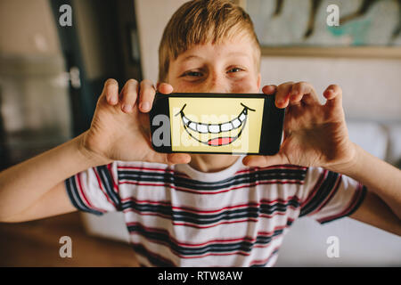 Boy holding un smartphone en face de son visage avec smiley photo sur l'écran. Garçon faisant semblant d'être heureuse à la maison. Banque D'Images