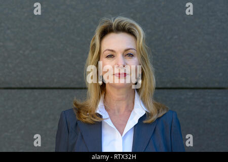 Jolie blonde femme professionnelle dans une élégante veste et chemisier blanc posant contre un mur gris smiling at the camera Banque D'Images