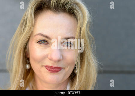 Close up face portrait d'une jolie femme blonde avec des yeux bleus et un sourire amical posant devant un mur gris Banque D'Images