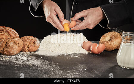 Baker casser des œufs dans un tas de farine à pâtisserie avec du lait dans un pichet qu'il prépare ses pains spéciaux Banque D'Images